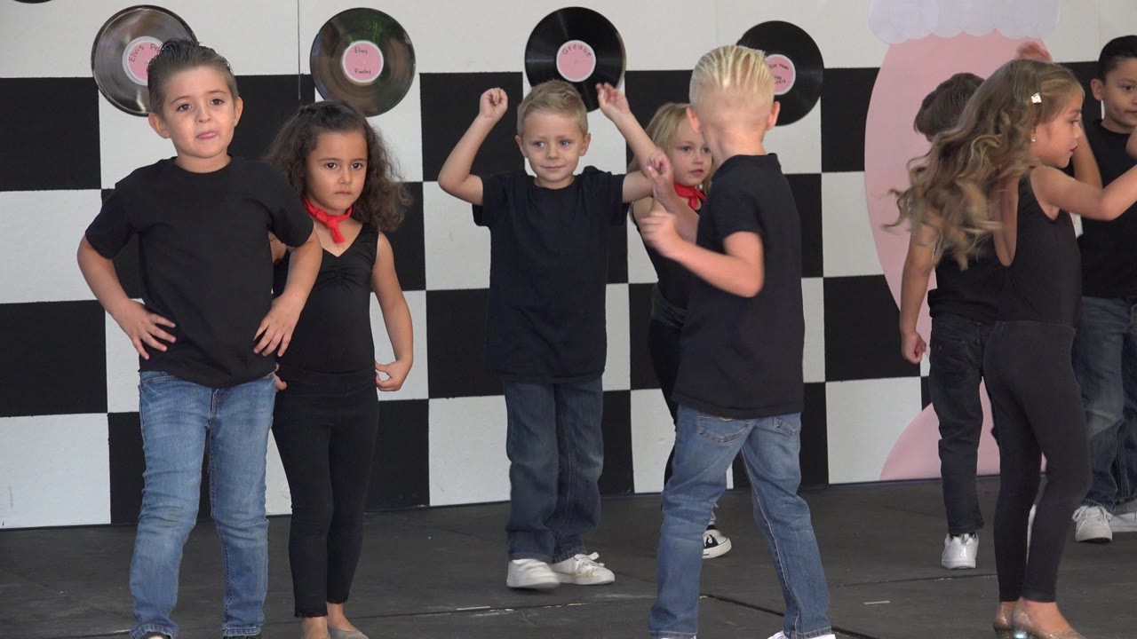 Children perform on a Cabo San Lucas stage in black outfits, with a boy standing hands-on-hips near the front and vinyl records on a checkered wall behind them.