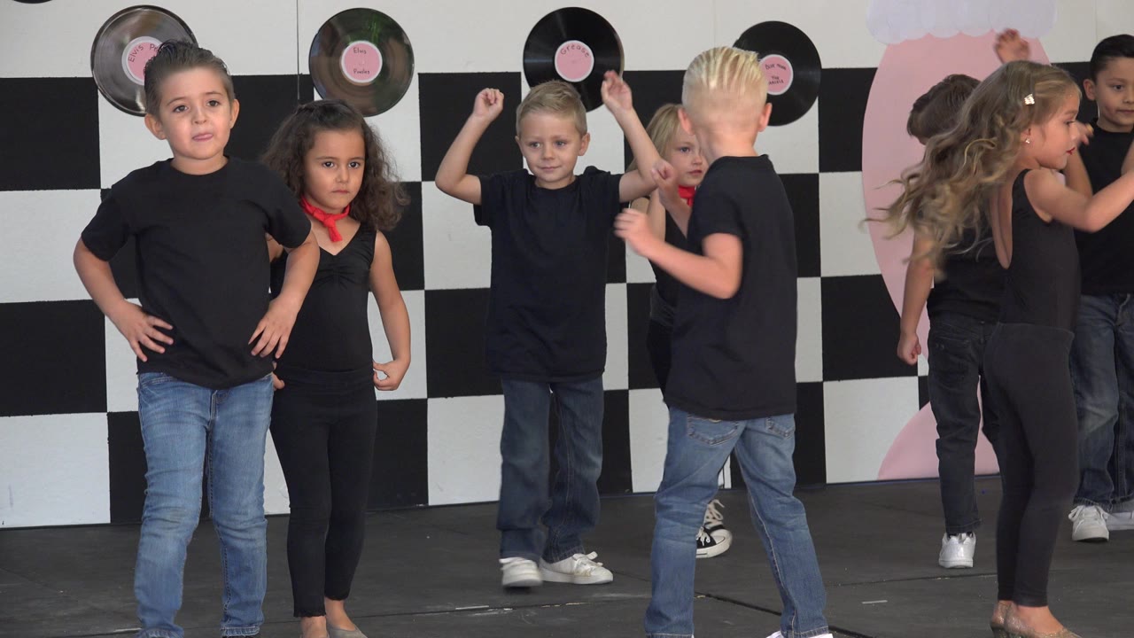Several children dance on the Cabo San Lucas stage, including a blond boy raising his arms and two girls moving near the pink-and-record backdrop.
