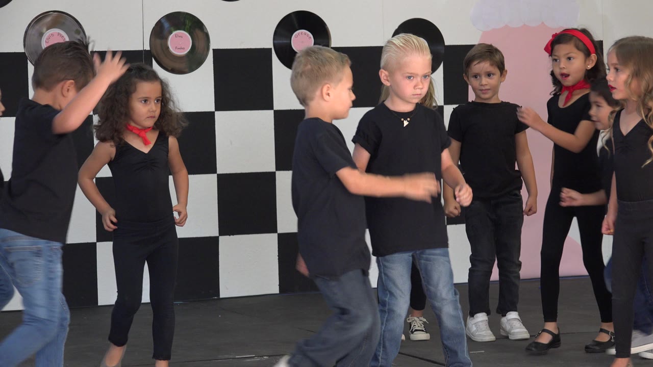 Several children in dark tops and jeans face inward during the dance on a Cabo San Lucas indoor stage, with black-and-white wall panels behind them.