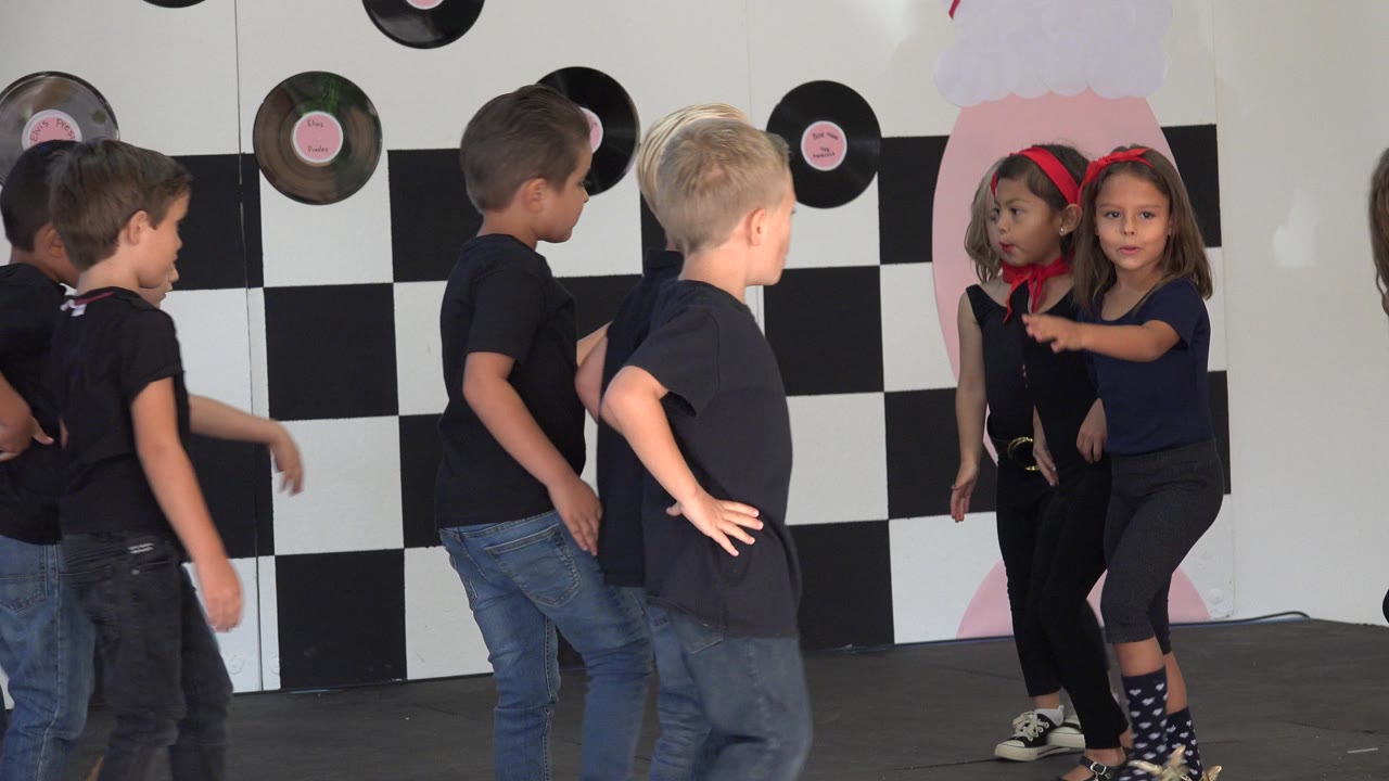 Children dance on a stage in Cabo San Lucas, Mexico, with boys at left and girls at right in front of a checkerboard wall and record decorations.