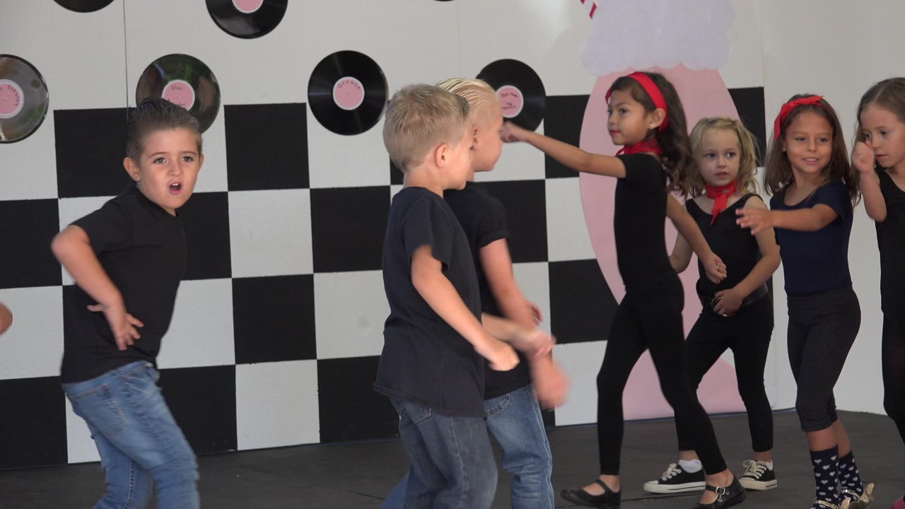 Children move across a stage as girls gesture toward the boys.