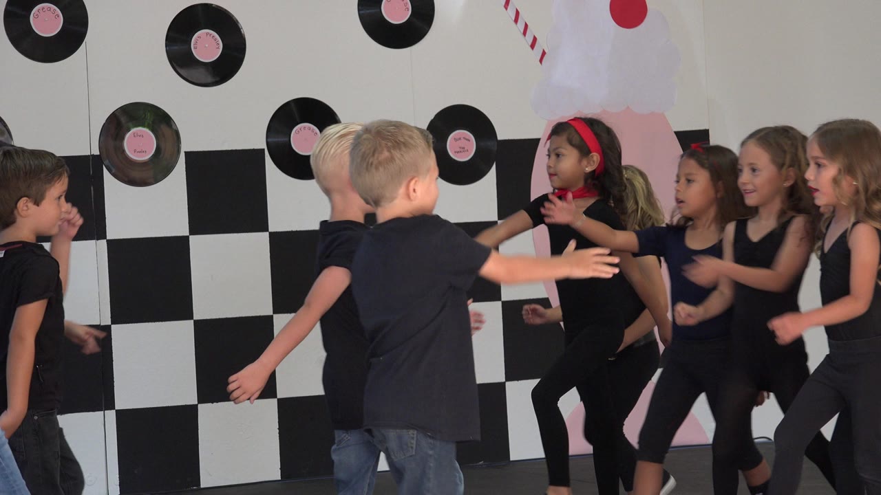 Children in black outfits dancing in front of a checkerboard stage backdrop.
