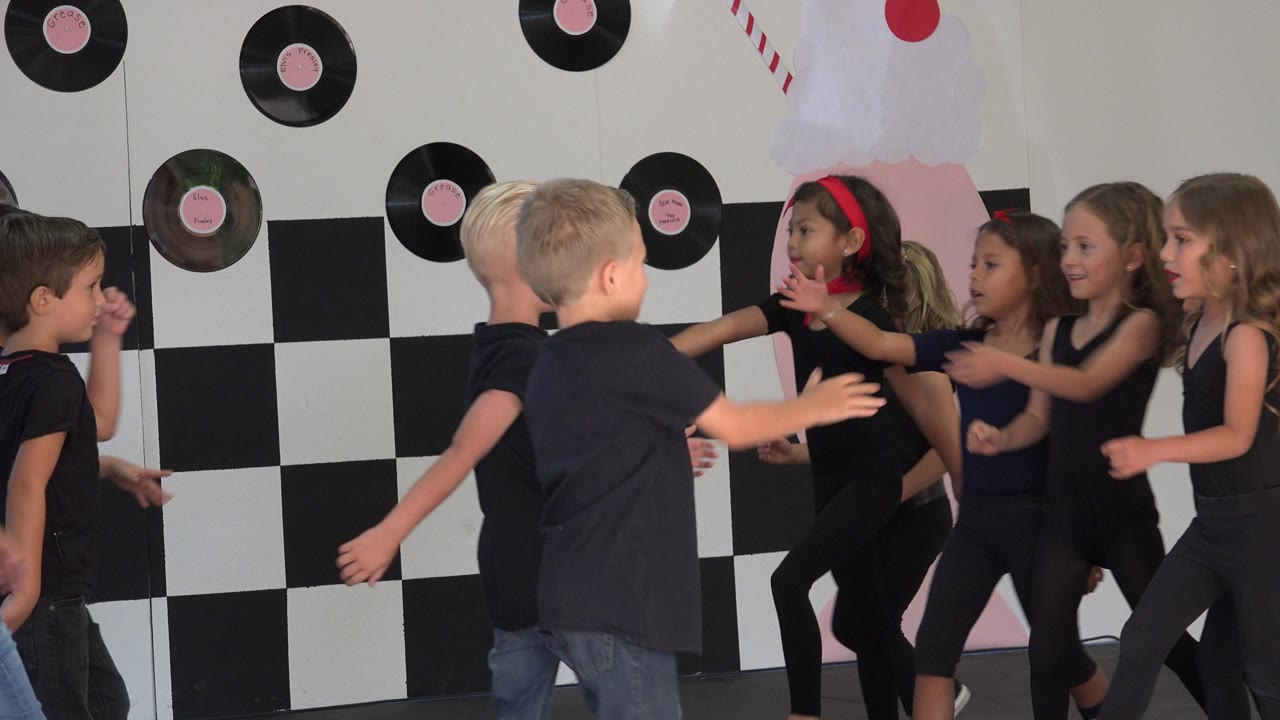 Several children in dark dance clothes move their arms and face one another on a Cabo San Lucas stage with record decorations and a checkerboard wall.