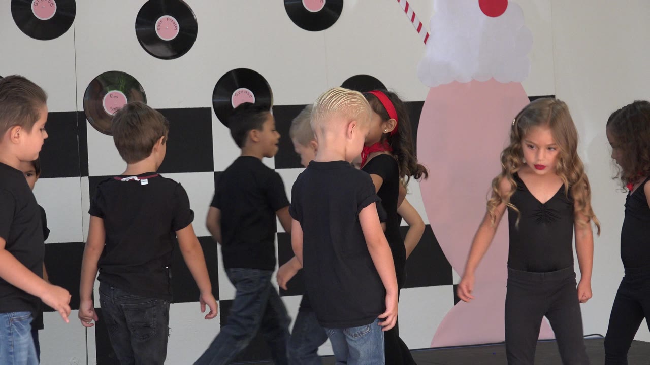 Children in black shirts and jeans stand and turn near a black-and-white checker backdrop with vinyl record decorations in Cabo San Lucas, Mexico.