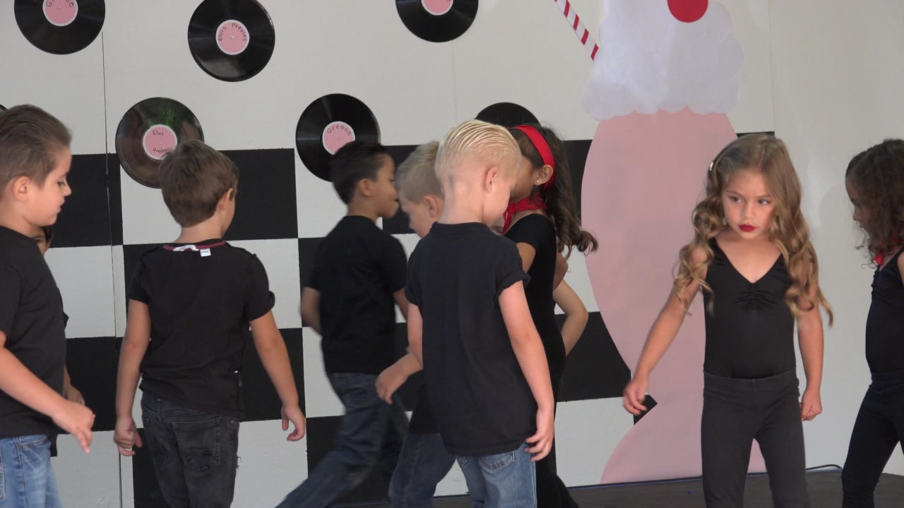 Several children move across the frame as a young girl in black stands near pink stage decor and vinyl records on the wall in Cabo San Lucas, Mexico.