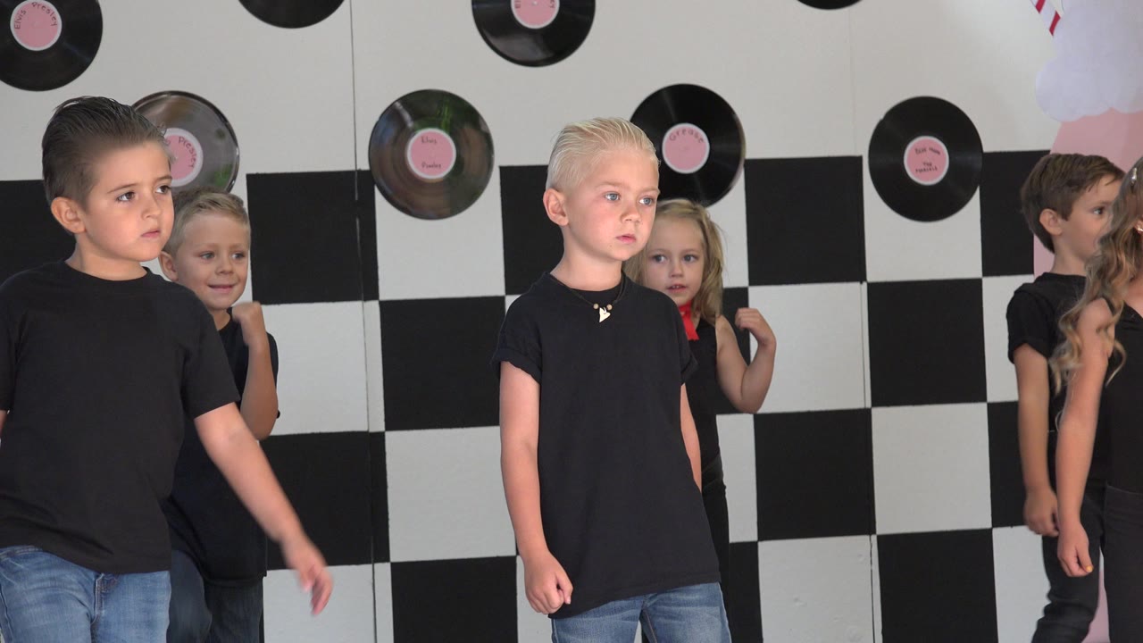 Children in black shirts walk across a checkerboard stage backdrop.