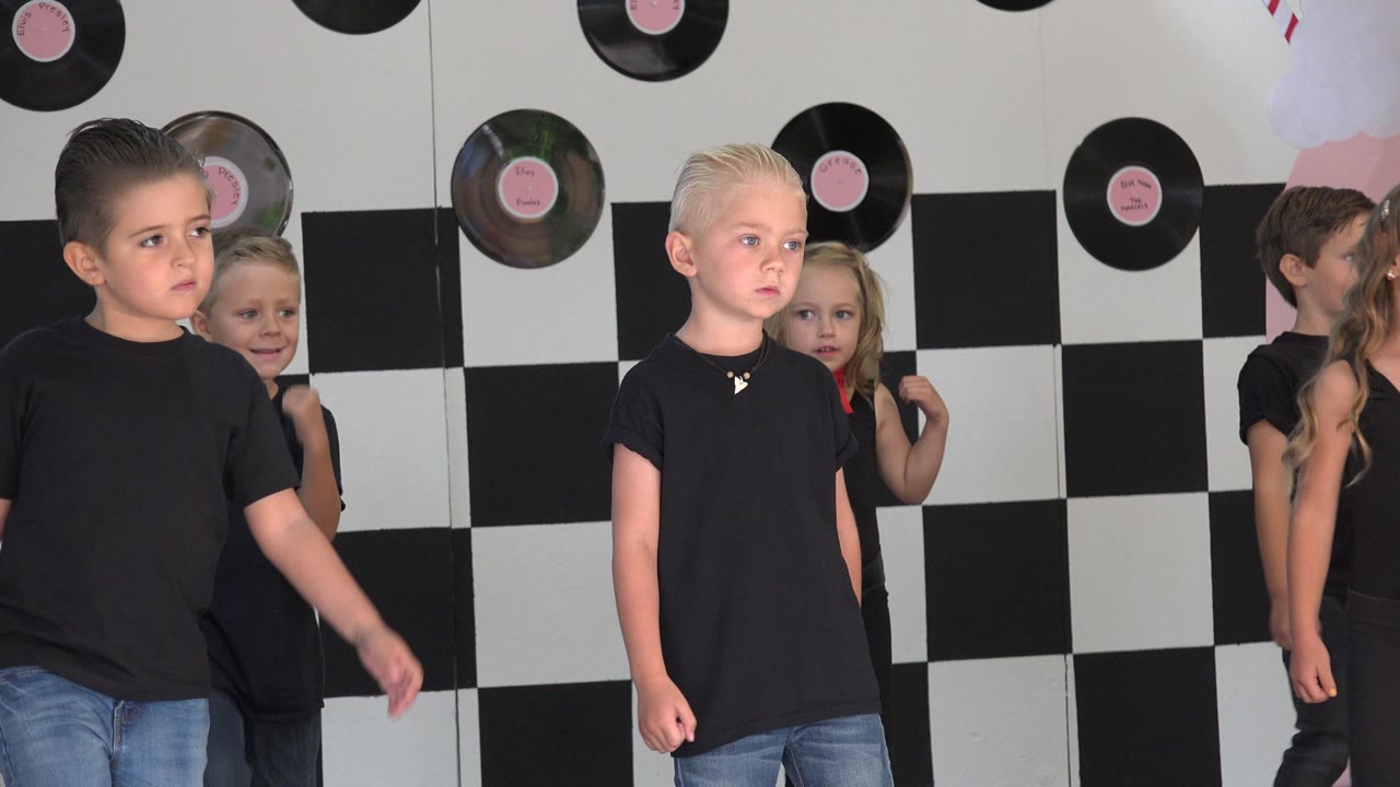 A blond child faces forward while other children gather behind him on the Cabo San Lucas performance stage with pink decor at the side.