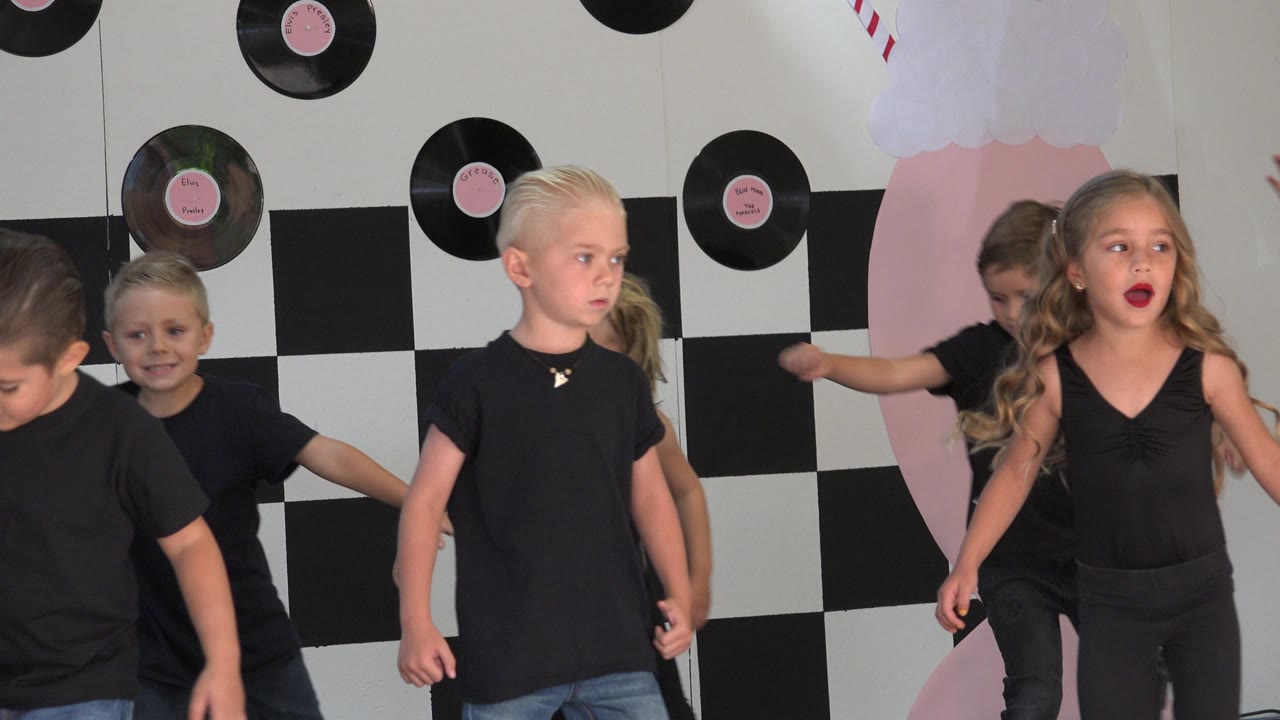 A blond child in a black shirt stands near center while a girl in black dancewear and other children move around him against the decorated Cabo San Lucas school stage wall.