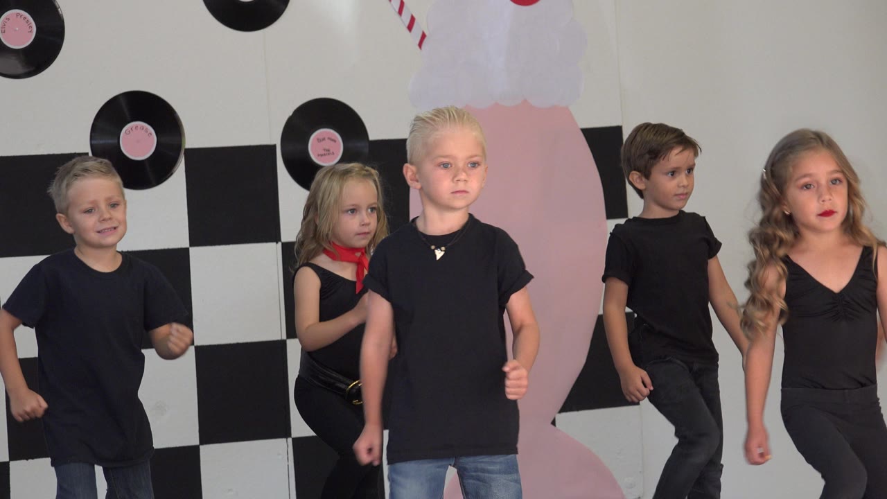 A line of children in black outfits continues the dance in Cabo San Lucas, Mexico, with a blond boy centered and record decorations on the wall.