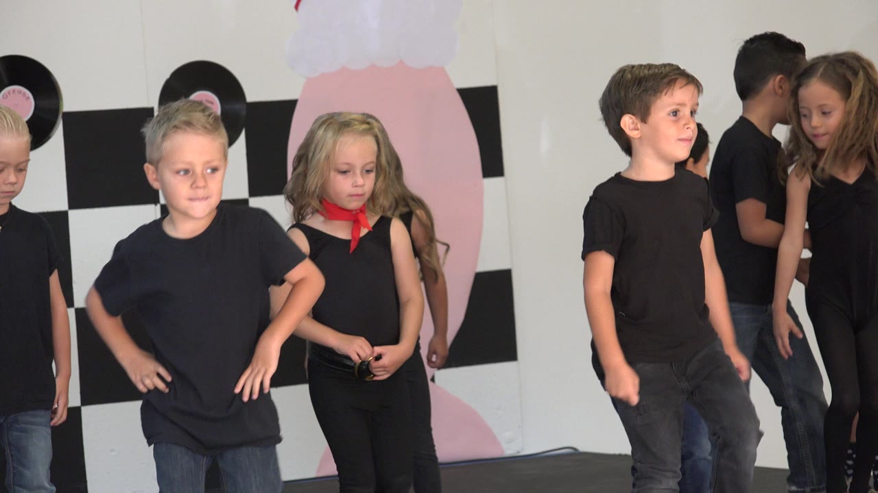 Children in black outfits stand on a decorated stage backdrop.