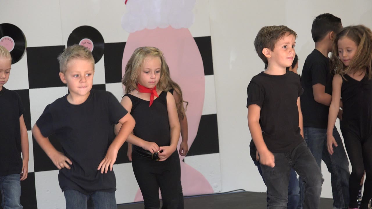At 0:01, a boy in a black shirt and jeans steps near the foreground as other children in black outfits wait behind him on the Cabo San Lucas stage set.