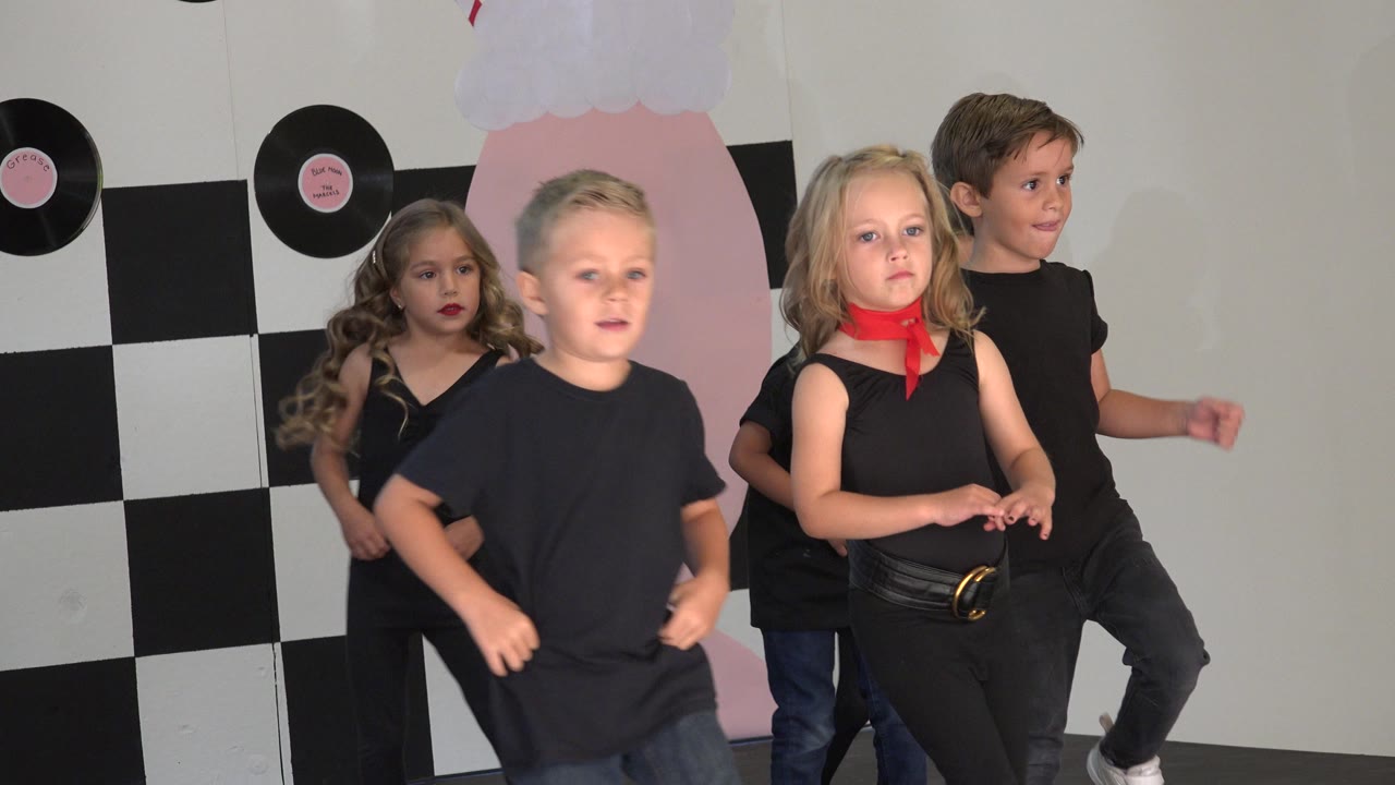 Young children in black dance outfits perform on a small Cabo San Lucas stage with a checkered backdrop, pink decor, and record decorations behind them.