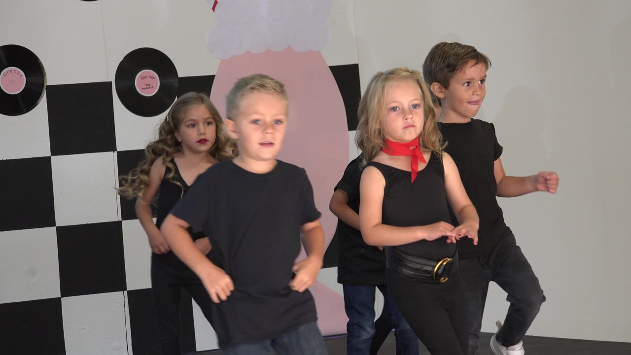 A blond boy moves near the foreground while other children in black outfits stand and dance near the checkered Cabo San Lucas stage backdrop.