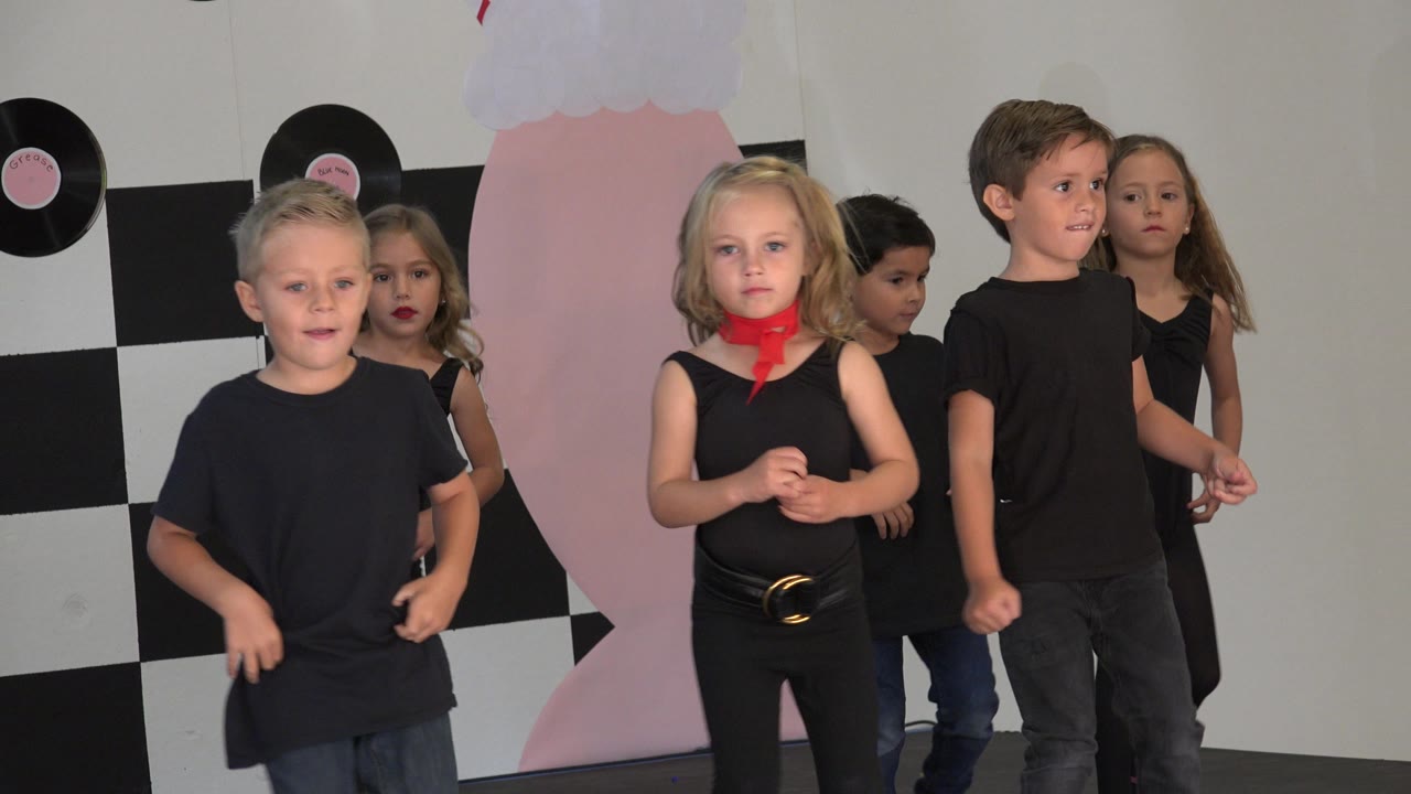 Children in black shirts and dancewear line up across the Cabo San Lucas stage, with the girl in a red scarf near center and checkered decor at left.
