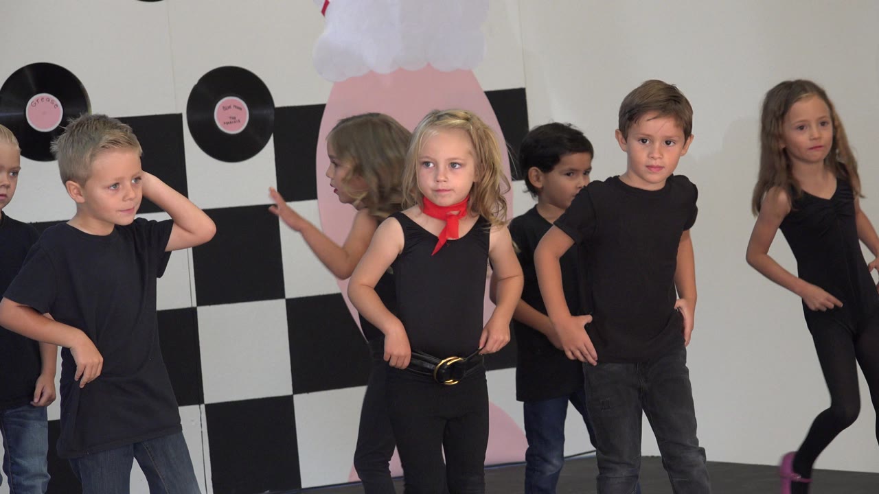 At 0:00 in Cabo San Lucas, Mexico, several children in black tops stand and dance in front of a checker-pattern stage backdrop with record decorations.