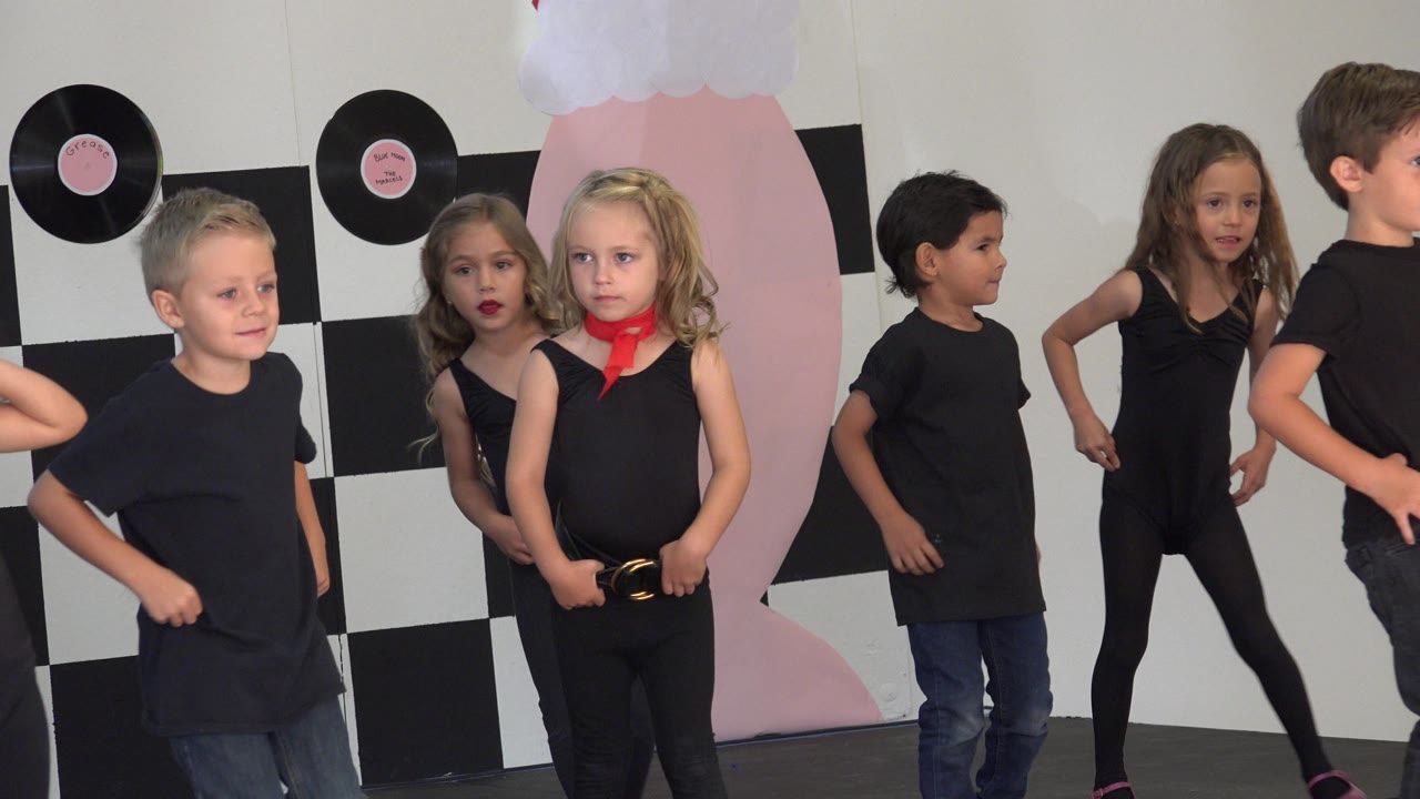 Young children in black outfits stand and dance in front of a checkerboard wall.