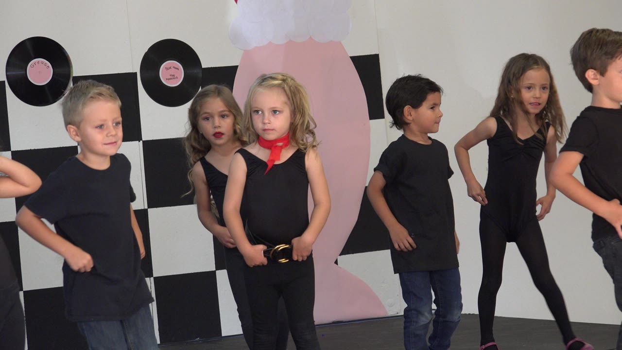 At 0:01, the blonde girl with the red scarf faces forward while other children in black shirts stand and move around her in the indoor Cabo San Lucas dance setting.
