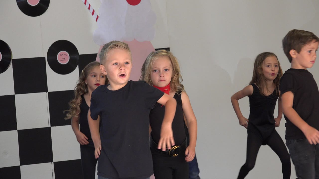 A boy in a black shirt and jeans stands to the right while girls in black dance outfits hold positions on the decorated stage in Cabo San Lucas, Mexico.