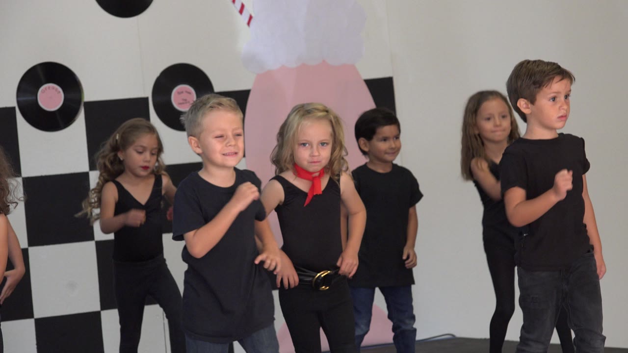 Children in black dance clothes stand before a checkerboard wall.