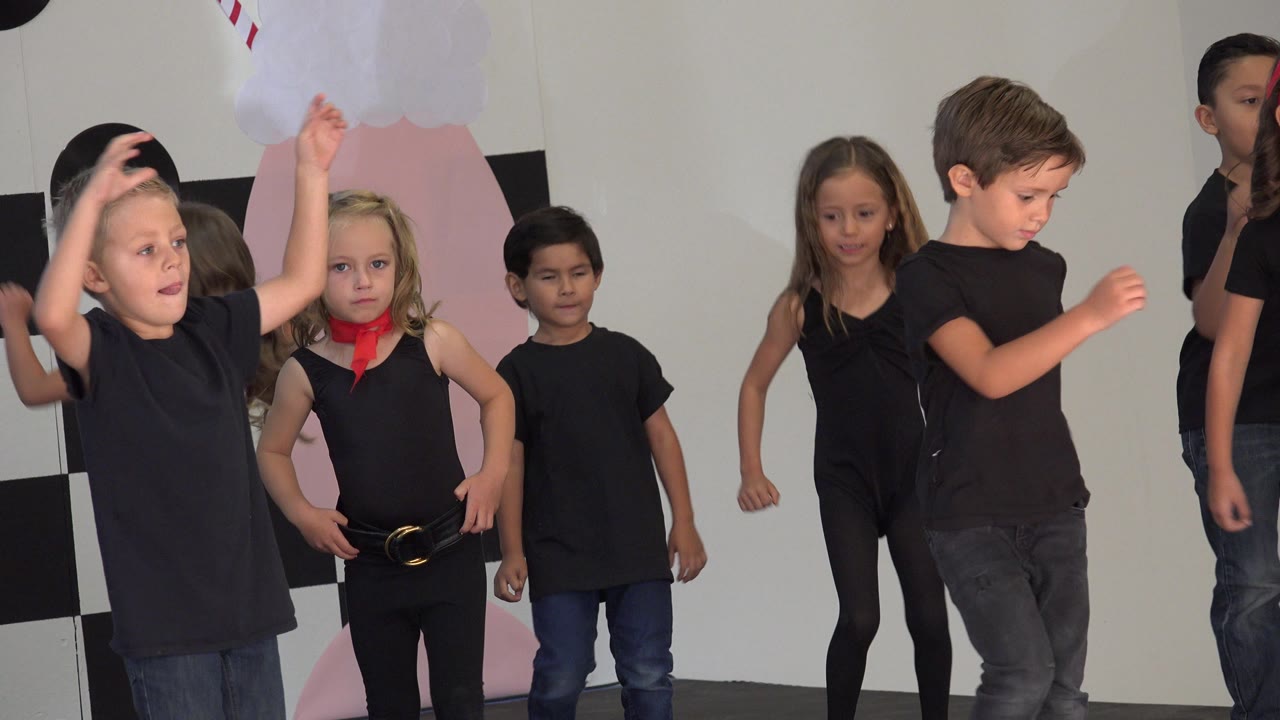 A boy near the front raises his arm while other children in black outfits continue the Cabo San Lucas indoor dance routine behind him.