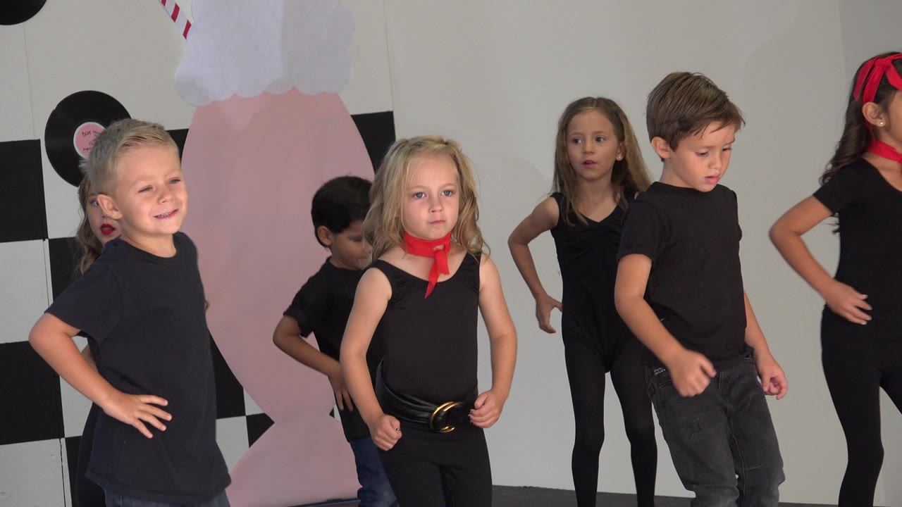 Young children in Cabo San Lucas stand in a loose line wearing black outfits, with red scarf accents visible on some dancers and a decorated wall behind them.