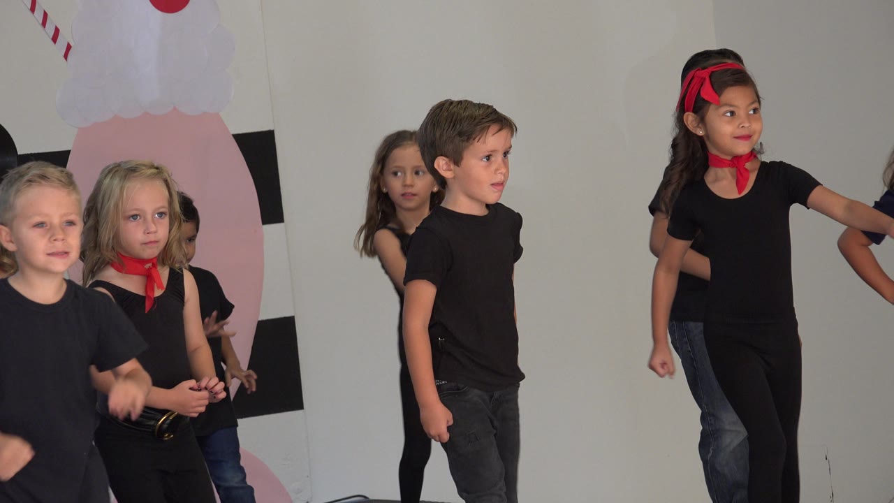 A boy in a black shirt gestures forward while other children stand behind him on the decorated indoor stage in Cabo San Lucas, Mexico.