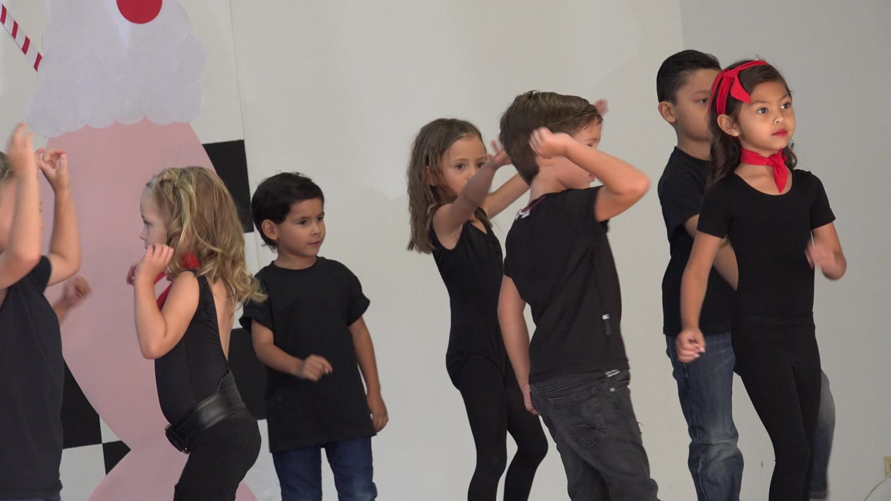 Young children in black clothing dance on a small decorated stage in Cabo San Lucas, Mexico, with a pink backdrop shape and record-style wall cutouts behind them.