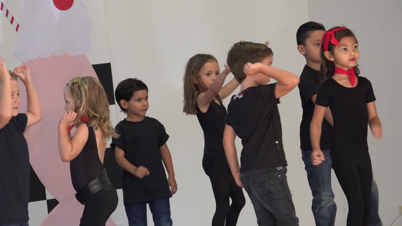 Children continue the stage routine in Cabo San Lucas, Mexico, with one boy raising an arm, a girl in a red headband facing forward, and others turning in black outfits.