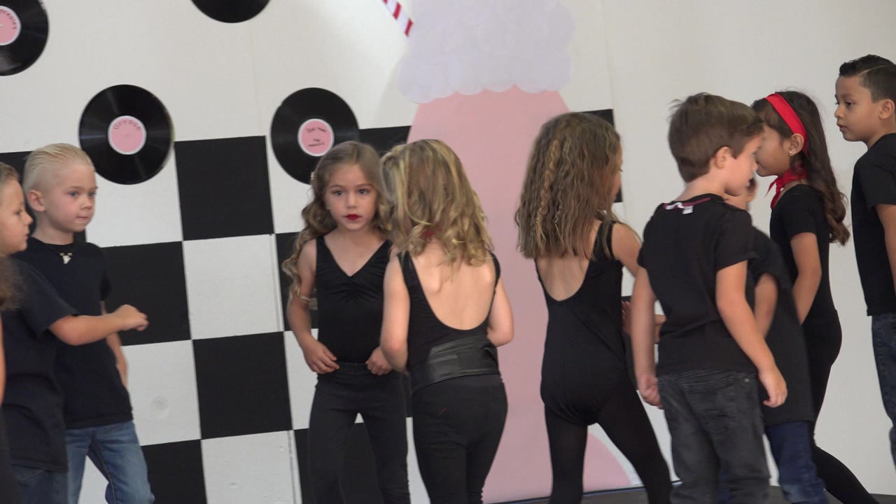 Young dancers in black clothing stand in a loose cluster in Cabo San Lucas, Mexico, with one child in a red headband near the right side of the frame.