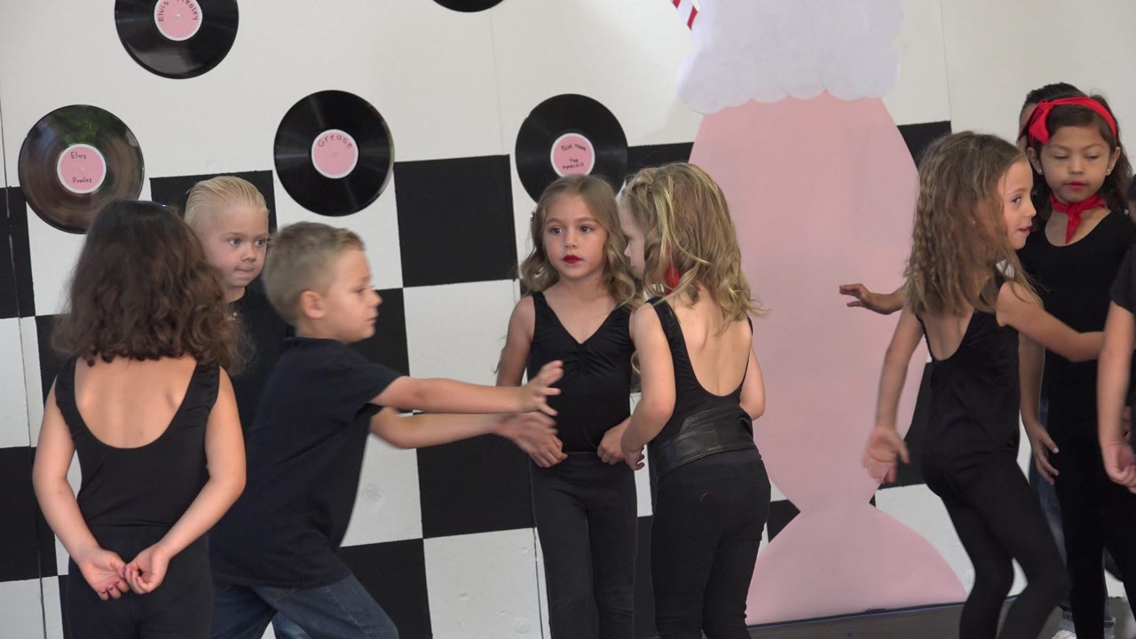 Children move and gesture near a pink stage prop and record decorations.