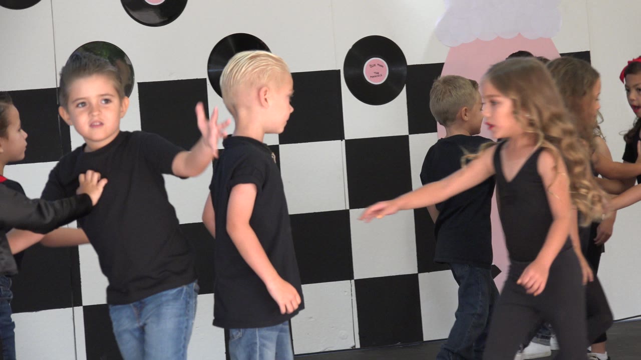 Children in black outfits dance in front of a checkerboard stage backdrop.