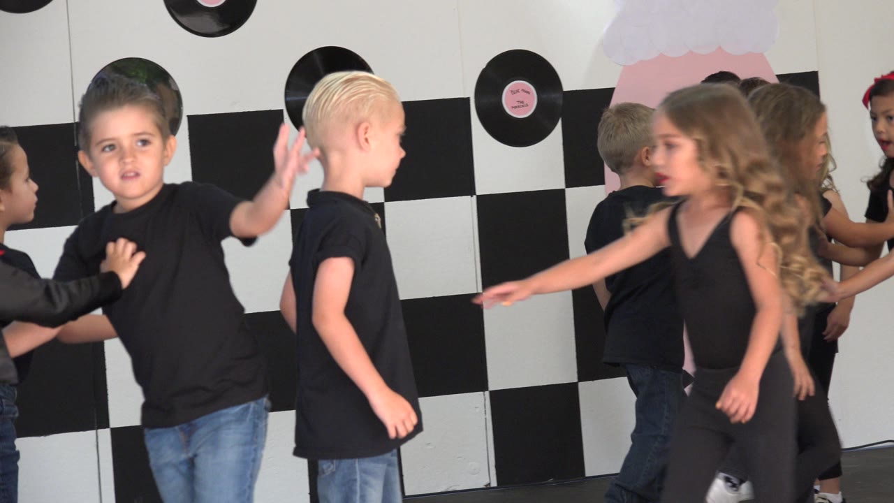 One second in, a blond boy and a girl with long hair pass near the camera while other children continue the routine against the decorated Cabo San Lucas stage backdrop.