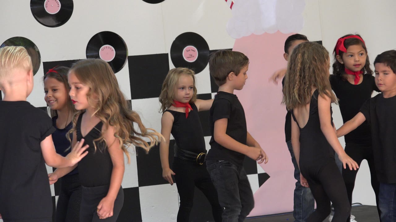 Young children in black performance outfits stand close together on a decorated stage in Cabo San Lucas, Mexico, with vinyl-record props and a checkered backdrop behind them.