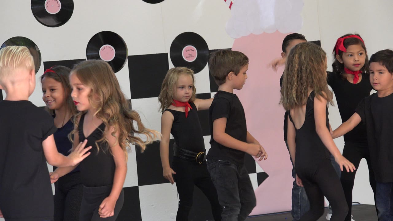 Children continue the routine on the Cabo San Lucas stage, with one girl facing forward near center and other children turning or reaching in front of the record-themed backdrop.
