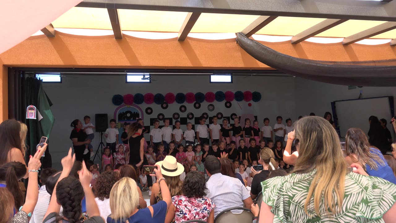 Wide audience view of a Cabo San Lucas school ceremony, with parents in the foreground holding phones and children grouped on a decorated stage.