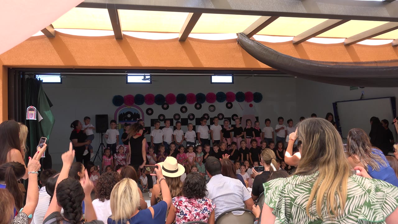 Audience members face a decorated indoor stage in Cabo San Lucas, where young children stand and sit in groups during the school ceremony.
