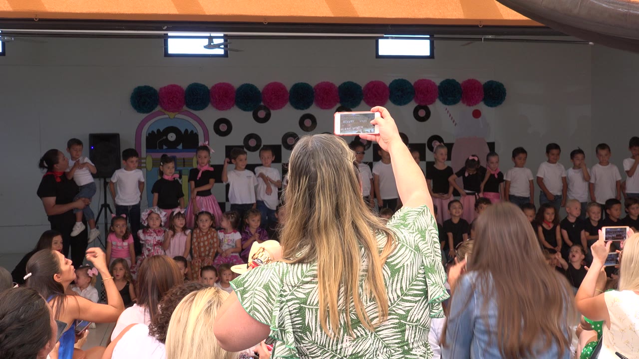 A woman in a green patterned shirt films over the audience as children stand and sit across the decorated school stage in Cabo San Lucas, Mexico.