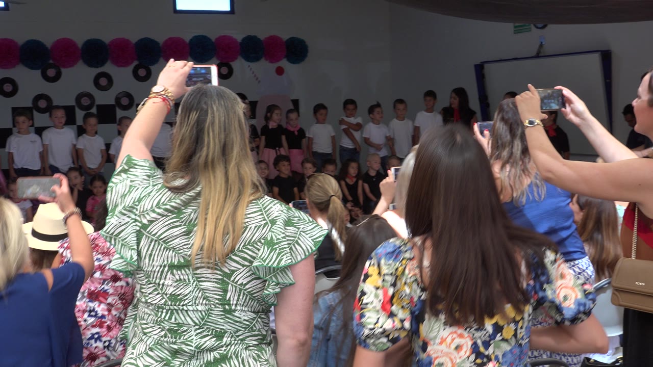 Several adults in patterned dresses and tops record children gathered along the rear wall of the Cabo San Lucas event room.