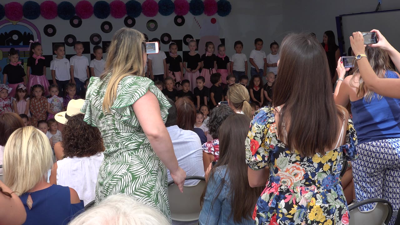 Audience members record children standing on a decorated indoor stage.