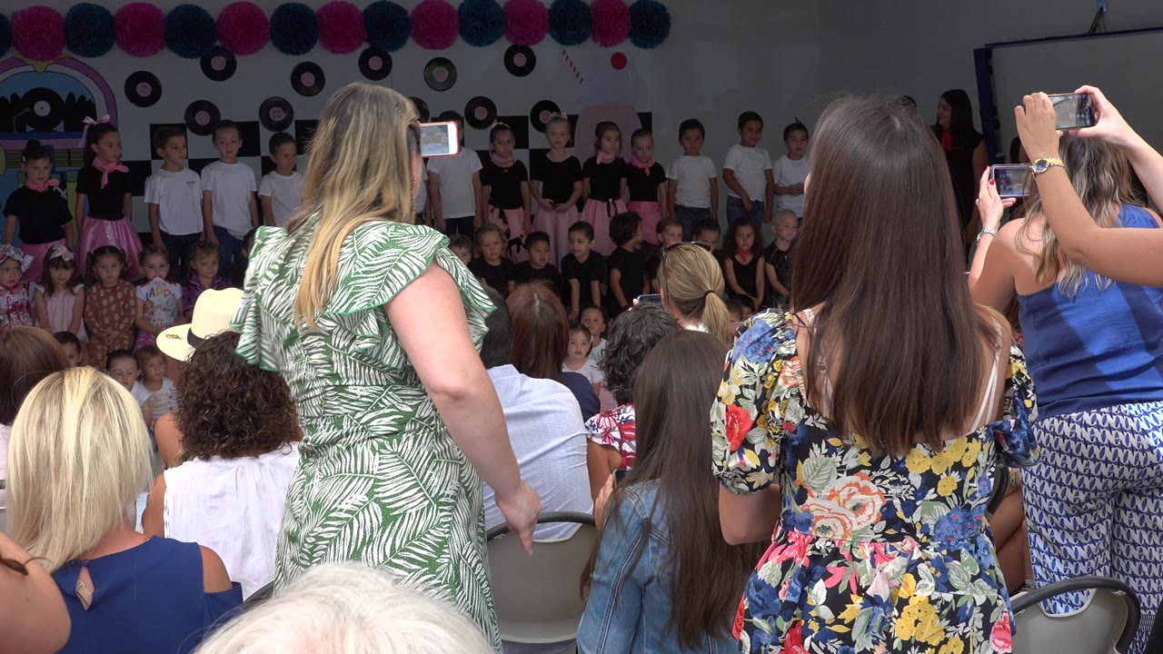 In Cabo San Lucas, Mexico, a woman in a green patterned dress stands near the center as other adults record the children lined across the stage.