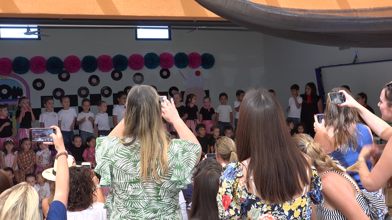 A woman in a green patterned top and other adults stand or sit in the foreground while children sing on a decorated stage in Cabo San Lucas, Mexico.