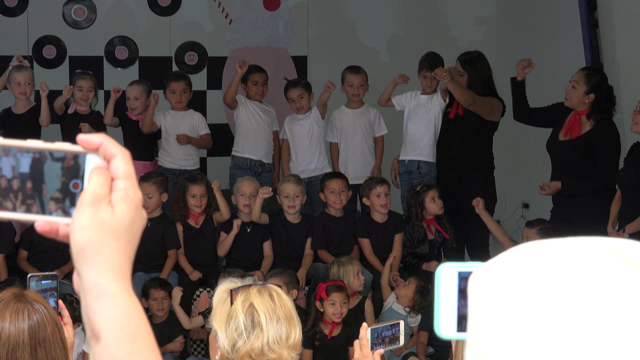 Children gathered on a small indoor stage while adults hold up phones.