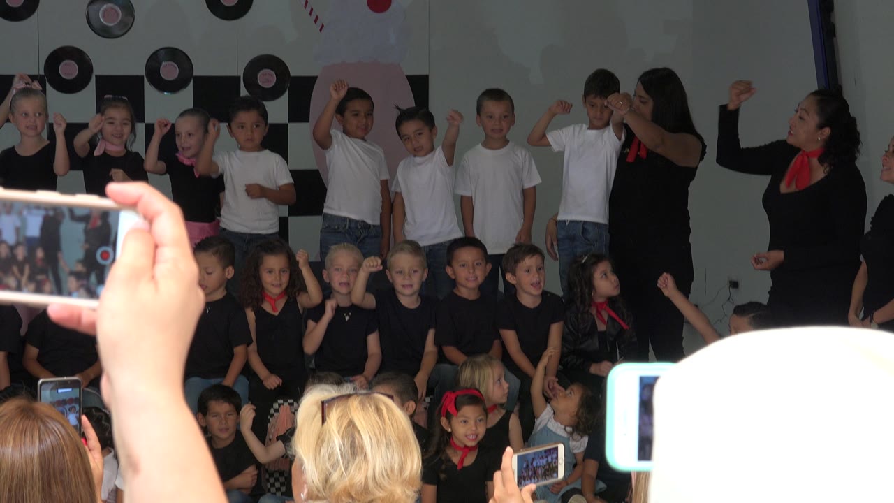 A wider audience view shows young children grouped on an indoor school stage in Cabo San Lucas, Mexico, with several phones visible in front.