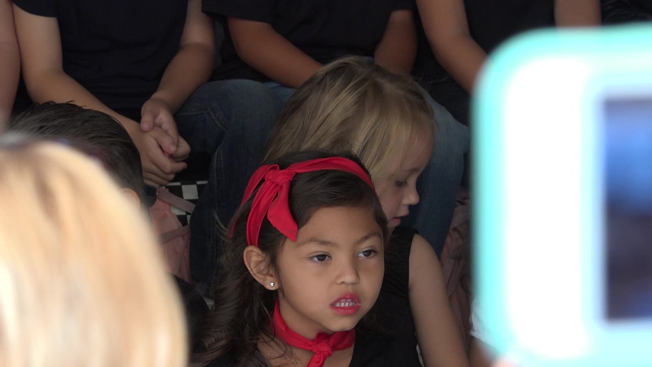 Young girl with red headband seated among children in an audience.