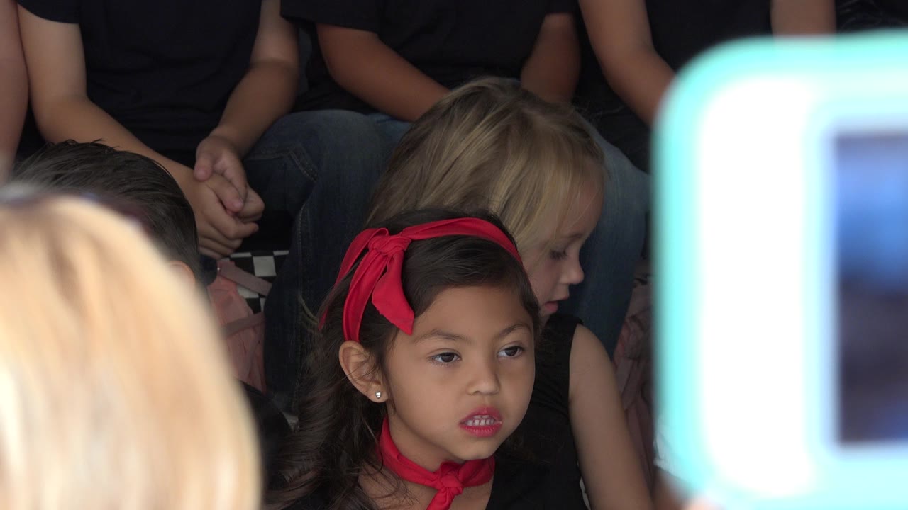 At 0:01 in Cabo San Lucas, the girl with the red headband remains centered, with a blonde child behind her and a bright out-of-focus phone screen along the right edge.