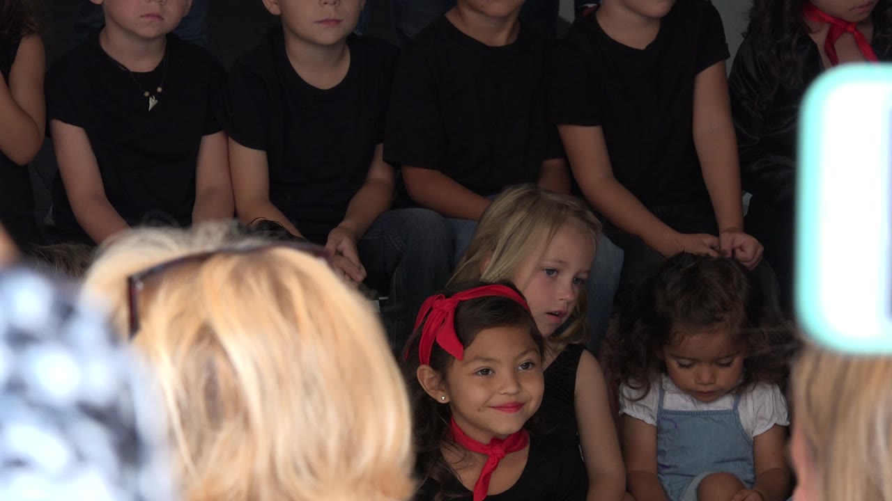 Children seated indoors with blurred adults and phone in the foreground.