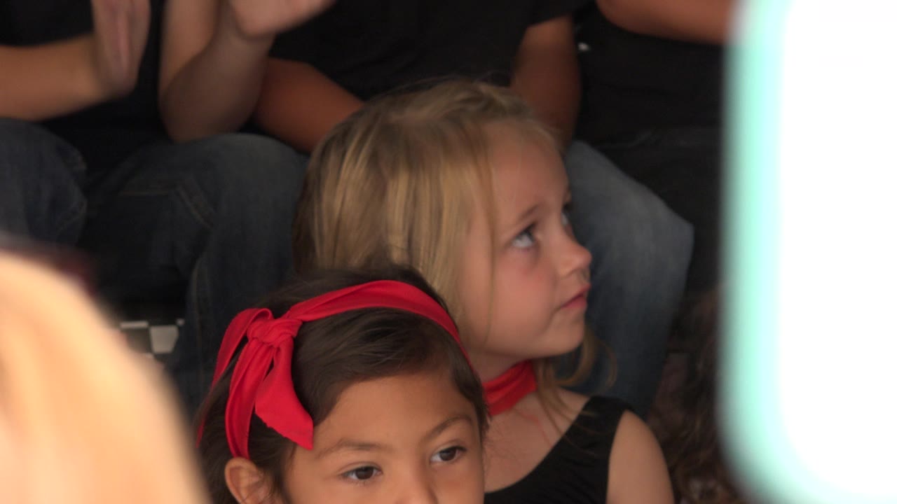Young girls seated in a crowded group, one wearing a red hair bow.
