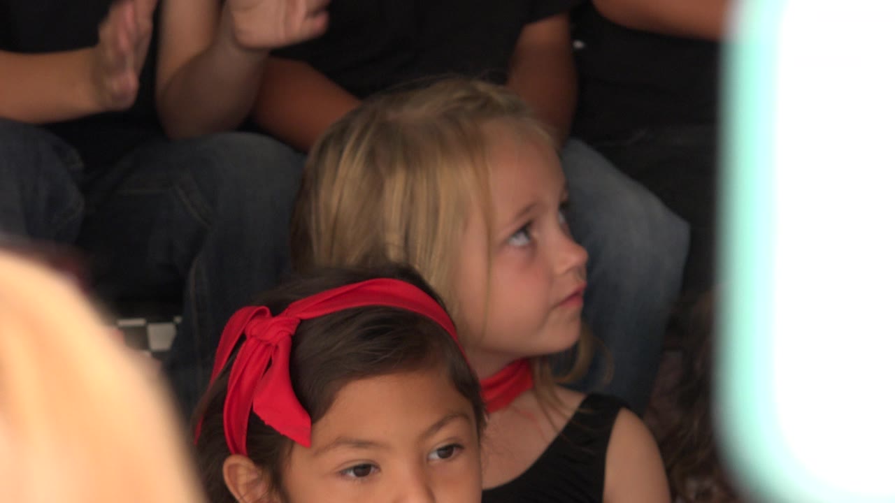 At 0:01, the blonde child remains seated and looks up toward something off frame while a girl with a red bow fills the lower foreground in the Cabo San Lucas crowd.