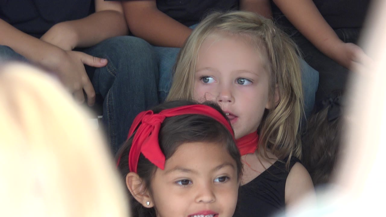 Children seated in dark shirts behind blurred audience heads.