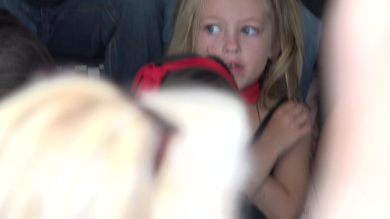 At 0:00 in Cabo San Lucas, a blonde child looks sideways while seated among other children, with bright blurred foreground arms partially blocking the view.